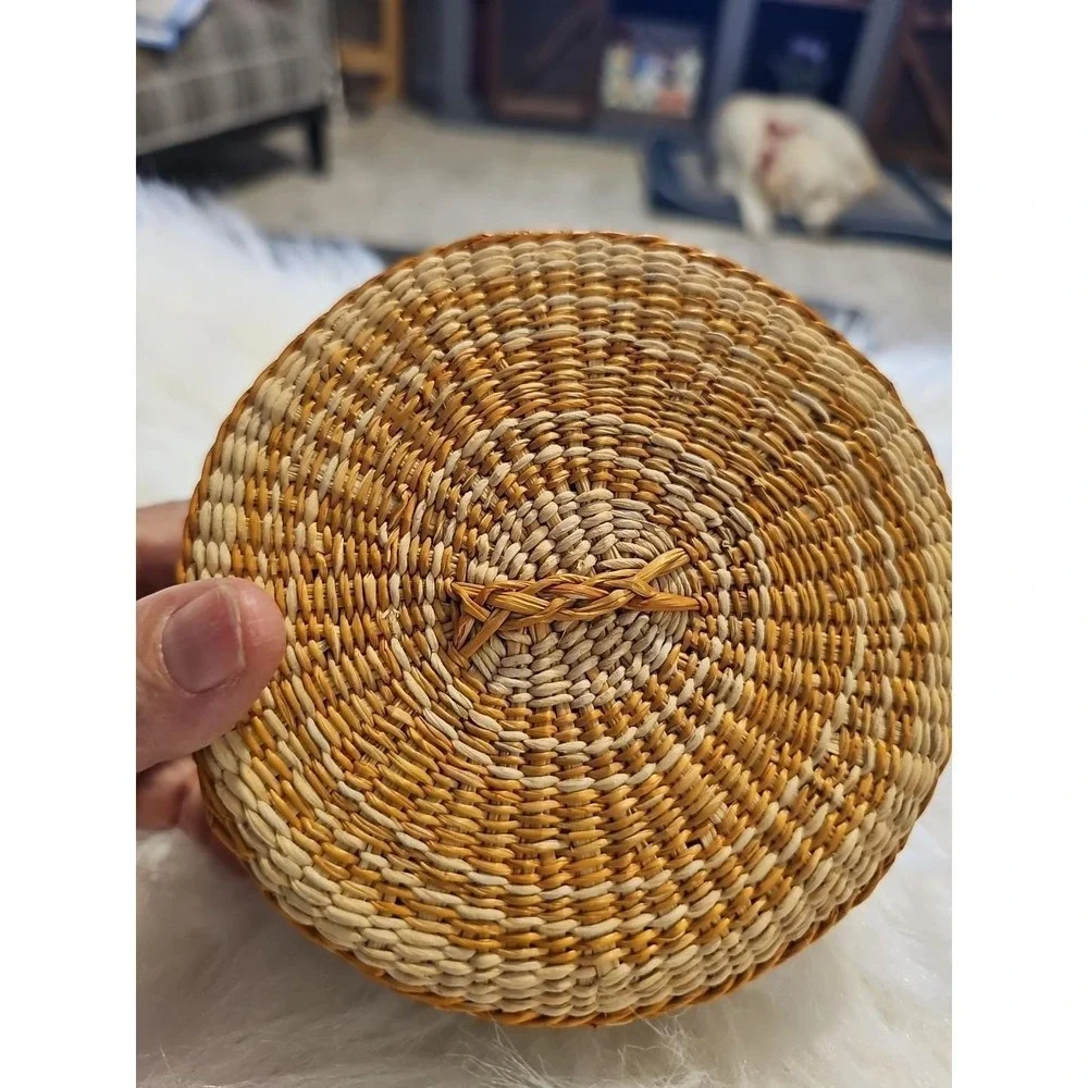 The image displays a‎ small, round, hand-woven basket with a lid, typically made